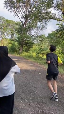The video shows two people from behind, walking on a paved road that goes through a wooded area. The person in the foreground wears a black hijab and a light blue and white shirt. The person in the background wears a black and yellow shirt and black shorts. The road is lined with trees and greenery. The sky is visible and appears to be partly cloudy.