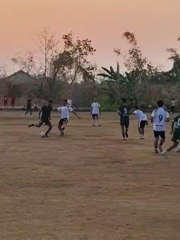 A group of young men are playing soccer on a dirt field. The sun is setting, casting a warm glow over the scene. The players are wearing casual athletic clothing, and some have numbers on their shirts. The field is surrounded by trees and vegetation, with a few buildings visible in the background. The game appears to be informal, with players scattered across the field and a ball in play. The overall atmosphere is relaxed and enjoyable.