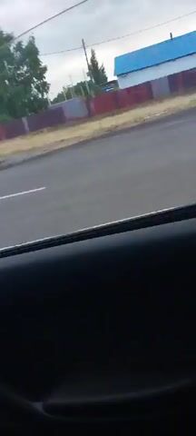 The video captures a view from inside a vehicle looking out a window. The scene outside is a roadside with a grey asphalt road with a white line. Beyond the road is a brown dirt area and a fence made of red and grey rectangular panels. Behind the fence are trees, utility poles, and buildings. One building has a white lower section and a bright blue roof. The sky is cloudy and grey. The video appears to be a short recording of a moving vehicle passing by this scenery.