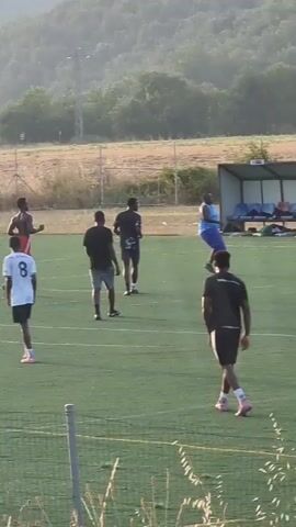 A group of six men are playing soccer on a green artificial turf field. The men are of varying ethnicities and build, with four visible in the foreground and two further back. One man in the foreground is wearing a white t-shirt with the number 8 on the back, black shorts, and pink and white cleats. Another man in the foreground is wearing a black t-shirt, black shorts, and pink cleats. In the background, a man in a white t-shirt and black shorts is running, and another man in a blue shirt and blue shorts is walking. A bench with a shelter behind it is visible in the distance, as is a fence and dry grass and trees. The lighting suggests it is daytime.