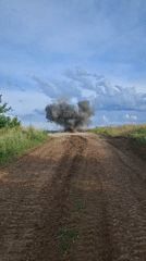 Dark smoke billows from a rural dirt road amid open fields