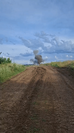 Dark smoke billows from a rural dirt road amid open fields