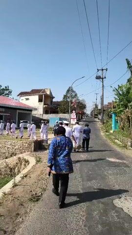 A group of people in white uniforms walks down a paved road. A person in a blue and gold patterned jacket and black pants walks away from the camera, also down the road. The road is lined with vegetation and buildings. The sky is clear and blue. The sun is shining.