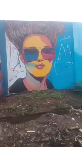The video displays a mural painted on a concrete wall. The mural is divided into several sections, each featuring different imagery. The leftmost section shows a colorful assortment of fruits, including what appears to be a grapefruit, pineapple, bananas, watermelon, and kiwi. To the right of the fruit section is a close-up of a woman's face wearing large sunglasses with colorful lenses. Her hair is styled in a voluminous updo. This section is painted against a bright blue background with some white graffiti-like strokes. The next section of the mural depicts two people at a market stall. One person, wearing a red hat, is interacting with another person wearing a blue shirt. The stall is laden with what appear to be bags of produce and baskets of fruit. Further to the right, another market scene unfolds, this time with three people behind stalls filled with various fruits and vegetables. One woman in a yellow top is arranging produce, while another person in a light blue shirt is also seen. The final section visible on the right shows another woman at a market stall, surrounded by baskets of fruit, including pineapples and what looks like mangoes. The ground in front of the wall is uneven and appears to be covered in dirt, rocks, and some debris, including plastic trash. A narrow ditch or channel runs across the foreground. The lighting suggests an overcast or diffused daylight.