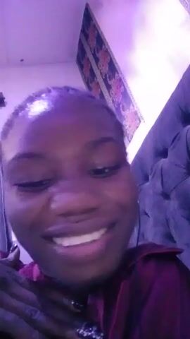 A young Black woman with close-cropped hair is the main subject of the video, which appears to be a selfie-style recording. She is wearing a dark red or maroon shirt and has a light, shiny complexion. She initially grins broadly, revealing her teeth, then sticks her tongue out playfully. Her expression shifts to a more neutral, slightly serious look as she looks directly at the camera. 

The background features a headboard of a bed in a dark color, possibly grey or black, and a decorative piece on the wall above. The room is bathed in a purple light, which affects the overall color cast of the video. Headphones are visible in her ears. The recording appears to be taken indoors.
