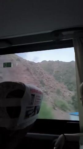 A first-person perspective video from inside a moving bus shows the view out the window. A person's head and shoulder are visible in the lower right corner of the frame, facing away from the camera. The landscape outside the window is mountainous with rocky terrain and sparse green vegetation. The bus appears to be traveling on a road that winds through the mountains. A sign with Chinese characters is visible on the back of a seat in front. The video is dark and grainy.