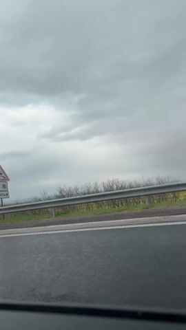 The camera is mounted inside a car moving along an asphalt road with a double white line dividing it. A guardrail separates the road from a bank of low, sparse vegetation. The sky is overcast with a dull gray. A road sign stands by the side of the road, displaying a warning about slippery conditions. Cars pass by, including a silver hatchback. As the car moves, a small, colorful card with cartoon images is held up in front of the camera lens, obscuring the view briefly. The focus is generally on the road and passing vehicles.
