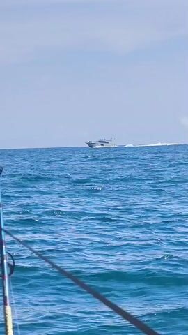 A fishing rod is positioned in the foreground of an ocean scene. The rod is blue with black accents, and it extends vertically and diagonally across the frame. In the background, a white yacht with a dark hull speeds across the blue water, leaving a white wake behind it. The sky is a clear light blue. The water is choppy with small waves.