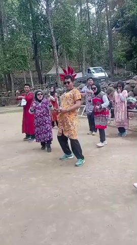 A group of eight people, five women and three men, are gathered outdoors in a dirt clearing surrounded by trees. They appear to be performing a coordinated dance or exercise routine. The person in the center, a man wearing a bright yellow and orange patterned tunic and black pants with green shoes, seems to be leading. He has a red headdress with a feathery top. To his left, a woman in a purple patterned dress with a black hijab is also dancing. To his right, a woman in a red and white patterned top and black pants with a grey hijab is moving. Other individuals are positioned around them, also engaged in similar movements. Some are wearing hijabs. In the background, there's a white vehicle, some rocks, and a small structure that might be a tent or shelter. The overall atmosphere seems festive or recreational.