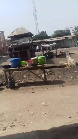 The video depicts an outdoor market setting with dirt and dust on the ground. There are several wooden tables with colorful buckets and other items on them. A bunch of sugarcane stalks lies near one of the tables. In the background, there are simple structures with corrugated iron roofs, one bearing the sign "AUDUMA OIL NIG. LTD". People can be seen walking around, and a few motorcycles are parked nearby. A tall communications tower is visible in the distance under a bright, slightly hazy sky.