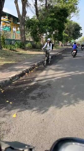 A person wearing a white cap, white long-sleeved shirt, and light-colored pants is riding a bicycle on an asphalt road. They have a backpack on their back. The bicycle is black with a silver frame. The person is in the middle of the road, with a sidewalk to their left. The sidewalk is lined with trees. In the background, there is a building with a yellow structure and green fencing. Further down the road, two motorcycles are visible, with one rider wearing a blue shirt and the other wearing a red shirt. The scene appears to be outdoors, during the daytime, with sunlight casting shadows on the road.