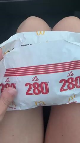 The video shows a person holding a McDonald's burger. The burger is wrapped in white paper with red and yellow branding. The burger itself is a long bun with a beef patty, cheese, tomato, and onion. The person is holding the burger in their lap, with their legs visible in the background. The lighting suggests it is daytime, possibly in a car. The burger appears to be part of a promotional item, indicated by the "McExtreme" branding on the wrapper. The person is unwrapping the burger and revealing its contents.