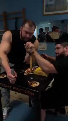 Two men are arm wrestling at a black table with the words "FIGHT FACTORY" on it. The man on the left is standing and wearing a black tank top, grey jeans and white sneakers, with a tattoo on his left arm. The man on the right is sitting in a black shirt. The man on the left is pushing down on the other man's arm, which is strapped to a yellow block. The room has blue walls, a wooden climbing apparatus, and a mirror with a Santa Claus decoration.
