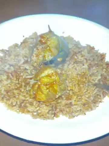 The video shows a white plate of what appears to be jollof rice with two pieces of fish on top. Steam is rising from the food, indicating it is freshly prepared. A spoon is partially visible, suggesting that the food is ready to be eaten. The background is simple and does not provide additional context. The camera focuses closely on the plate, emphasizing the dish. A blue and black charging cable is visible on the table.