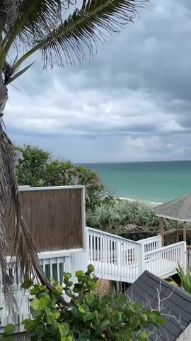 This is a vertical video of a cloudy sky over a turquoise ocean. In the foreground, there is a white wooden deck with railings and stairs, and a small hexagonal gazebo. Lush green vegetation and palm trees are visible around the deck and gazebo. The tide is out, revealing a sandy beach. The sky is filled with dark, heavy clouds, suggesting an impending storm. The camera is positioned at a high angle, looking down and out towards the ocean.