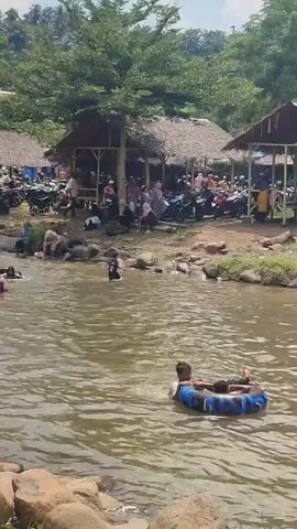 The video shows a river with many people swimming and playing in the water. There are several huts with thatched roofs along the riverbank, and many motorcycles are parked in front of them. The area is surrounded by lush green trees and vegetation. The water is murky brown and appears shallow in some areas. Several people are using inflatable tubes to float in the water. Children are visible among the swimmers. The overall scene suggests a recreational area or a tourist spot where people are enjoying a day out by the river. The sky is partly cloudy.