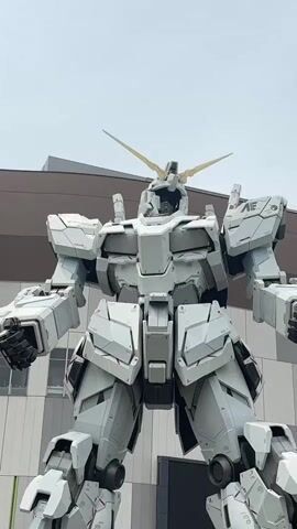 The video shows a full shot of a giant white robot from the chest up, with its arms held down by its sides. The robot has a horn on its head that glows yellow. The background is a cloudy sky with a modern building behind the robot. The robot's head is tilted slightly down. Its horns are angled upwards.