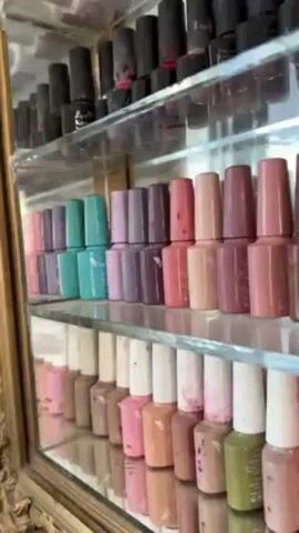 The video shows a display cabinet filled with rows of nail polish bottles in various colors. The cabinet has glass shelves and an ornate, gold-colored frame. The top shelves hold primarily dark-colored polishes, including black and dark grey. The lower shelves contain a range of lighter colors such as pink, purple, brown, green, red, yellow and blue. The camera pans from left to right, showcasing the variety of colors and the arrangement of the bottles. The backdrop is slightly blurred, suggesting the cabinet is located in a shop or salon setting.
