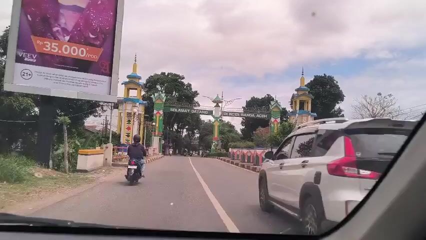 The view from inside a car shows a road leading through a colorful archway marking the entrance to Banda Aceh, Indonesia. The archway is decorated with green, yellow, blue, and white paint.  The words “SELAMAT DATANG DI KOTA BANDA ACEH” (Welcome to the City of Banda Aceh) are written across the archway. Two motorcycles and a white SUV are seen driving through the archway. There are buildings, trees, and light poles on either side of the road. The sky is cloudy.  A billboard is visible to the left of the road, showing a price of 35,000 per POD. The shot is stable and the focus is on the road ahead.