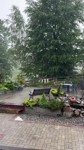 The video shows a patio area during a heavy rainstorm. The patio is paved with light-colored bricks and has several decorative elements, including a black metal bench, sculpted evergreen bushes, a pile of rocks with decorative pottery, and some old wooden wagon wheels. Beyond the patio is a grassy area with trees, a wooden fence, and a building with a red roof and siding. The rain is falling heavily, creating visible streaks of water in the air and causing the ground and objects to appear wet. The overall atmosphere is one of a stormy, wet outdoor environment.