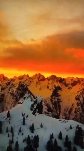The video shows a landscape of snow-covered mountains. The sky above the mountains is a blend of orange and yellow hues, indicating either a sunrise or sunset. Scattered evergreen trees dot the lower, snow-covered slopes of the mountains.