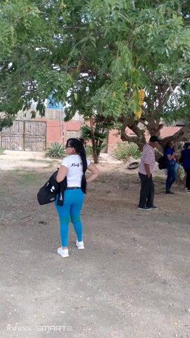 Several individuals stand or walk on a dirt path under the shade of a large tree. A woman in blue leggings and a white shirt walks toward the camera, while others congregate further down the path. A man in a white shirt and khaki pants walks in front of the camera. Some people appear to be taking pictures or looking at something further down the path. The scene is outdoors, with a building partially visible in the background.
