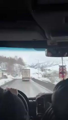 The video is taken from inside a vehicle driving on a snowy road. A white truck is driving in front of the vehicle. Several people are walking on the side of the road next to the truck. In the background are snowy hills and mountains. The interior of the vehicle is visible. A Georgian flag is hanging from the rearview mirror. The driver's hands are visible on the steering wheel. The faces of passengers are visible in the rearview mirror.