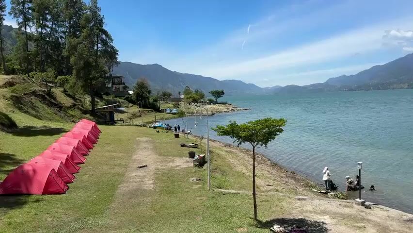 The video opens with a panoramic view of a serene lake with mountains in the background under a clear blue sky with scattered clouds. The camera pans from left to right, revealing a campsite situated on the grassy shore. In the foreground, a row of bright red tents is neatly arranged on the grass. Further into the scene, along the lake's edge, several people are visible. Some appear to be standing near the water, and others are seated, possibly enjoying the view. A small tree stands prominently on the shoreline. As the camera continues its pan, the view shifts to reveal more of the lake and the distant mountains. More tents, this time orange, are visible in the right portion of the frame. The water is calm and a vibrant turquoise color, reflecting the sky. The environment appears to be a popular spot for camping and outdoor recreation.