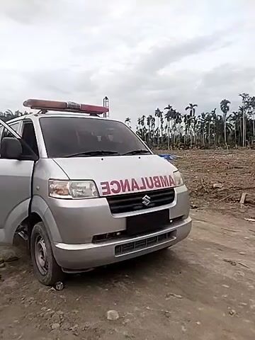 The video shows a silver ambulance parked on a dirt road next to a cleared field. The ambulance has the word "AMBULANCE" written in red letters backward across the hood. A man walks past the front of the ambulance, looking at the camera. The sky is overcast, and there are palm trees in the background.