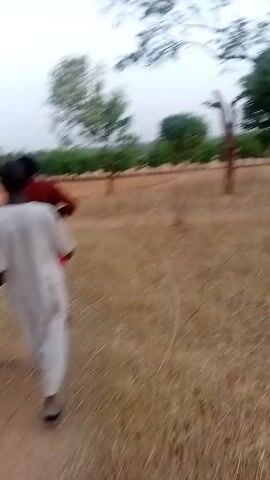The video shows a person walking through a field with sparse trees. The ground is covered in dry grass and reddish soil. Two people are briefly visible, one wearing a white shirt and holding a stick, and another in a red shirt. The sky is overcast. The camera movement is shaky, indicating it is handheld.