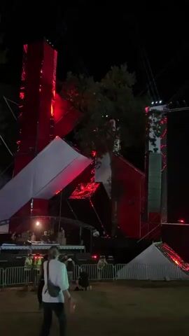 At night, a diverse group of people are gathered outdoors near a stage. The stage has a large, abstract design with red and white geometric shapes and is illuminated by red and blue lights. There are trees behind the stage. Several people appear to be working on the stage. In the foreground, three people are visible from behind. One person is walking away from the camera. Another person is standing with their back to the camera, and a third person is walking toward the camera. There are metal fences in front of the stage.