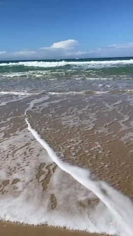 A serene beach scene unfolds with gentle waves washing over the sandy shore under a clear blue sky. The ocean's surface is a mosaic of turquoise and white, with waves creating foamy patterns as they recede. Sunlight glints off the wet sand, highlighting the texture and footprints left by unseen visitors. A few wispy clouds dot the horizon, adding depth to the vast expanse of the sky. The sound of the ocean is likely a calming murmur, accompanying the visual tranquility.
