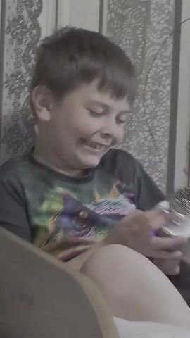 A young boy with short, light brown hair is captured in a close-up shot indoors. He appears to be between 5 and 8 years old. He is wearing a dark t-shirt, part of which shows a green and yellow pattern, possibly a dinosaur graphic. The boy is holding a silver-colored microphone with a textured, metallic head and a smooth handle. A subtle blue or purple light emanates from a section of the microphone's body, suggesting it might be an illuminated toy or karaoke microphone.

The boy is actively engaged with the microphone, holding it close to his mouth. He is laughing heartily, with a wide-open mouth, bright eyes, and a joyful expression that suggests extreme happiness or excitement. He is seated, possibly on a light-colored chair or sofa, with his bare legs visible.

The background consists of a light-toned wall decorated with an intricate, lace-like or floral pattern, possibly wallpaper, indicating a home setting. The lighting appears soft and natural, suggesting it is daytime. The overall ambiance is cheerful and lively, centered around the boy's joyful interaction with the microphone. While some text and a logo are visible on the microphone's handle, they are not clearly legible due to the resolution and angle. The event takes place in Kalipetrovo, Bulgaria.