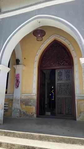The video shows the entrance to a building with an arched doorway. The doorway is made of dark wood and features intricate carvings. The walls of the building are painted yellow and appear weathered, with some peeling paint. Above the doorway, a decorative wicker lantern hangs. 

Inside the doorway, a person in a blue uniform with a yellow sash is visible, standing near a red rope barrier. Further inside, stained glass windows with blue and yellow panes can be seen. The overall scene suggests a historical or religious building, possibly a church or a colonial-era structure, given the architectural style and the presence of a uniformed individual, potentially a guard or attendant. The lighting suggests it is daytime. The location is identified as Hội Tín, Vietnam.