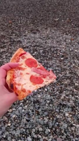 The video shows a slice of pizza held in someone's hand against a backdrop of a pebble beach and ocean waves. The sky is cloudy with hints of pink and orange from the sunset. The pizza slice has pepperoni and cheese. The waves crash onto the dark pebble-covered beach. The scene evokes a sense of relaxation and enjoyment of food in a natural setting.