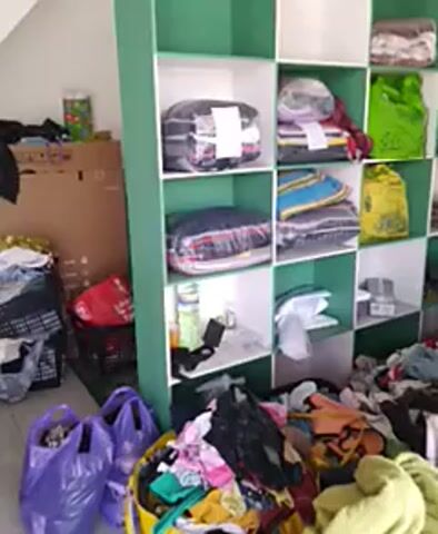 The video shows a cluttered storage area. There are shelves filled with neatly folded items, some wrapped in plastic. To the right, the shelves are overflowing with piles of clothes. Below the shelves, there are more piles of clothes and plastic bags. The area appears to be indoors, with white walls and green shelving. The lighting is somewhat dim.