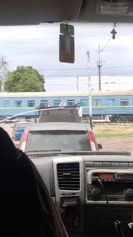 The video provides a first-person view from inside a vehicle, looking out through the windshield. Several cars are stationary in front, suggesting a queue or a stop at a railway crossing. In the background, a blue passenger train, consisting of multiple carriages, is visible on the tracks, either halted or moving slowly. A white sign with the number "45" is situated near the railway line. The scene is set outdoors during the daytime, under an overcast sky. Inside the car, the dashboard is visible, featuring a digital clock displaying "19:37" and a car radio unit. An air freshener hangs from the rearview mirror.