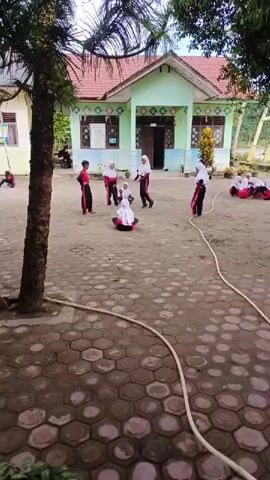 The scene shows a group of children, mostly girls, dressed in school uniforms with hijabs, playing in a schoolyard. The school building is in the background, with a light green facade and a red tile roof.  The yard has a hexagonal tiled surface. A long, white hose lies across the yard. Some children are standing, some are sitting on the ground, and they appear to be engaged in some sort of game. A tall, slender palm tree is visible on the left side of the frame. The overall impression is that it's an ordinary day at an Indonesian school, with children enjoying their time outside.