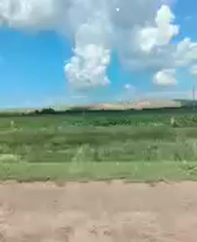 The video shows a landscape with a dirt road in the foreground. Beyond the road is a green field with some scattered trees or structures. In the distance, there is a ridgeline or hills under a blue sky with white clouds. The camera appears to be moving, suggesting the footage was taken from a vehicle. The scene is rural and appears to be daytime.