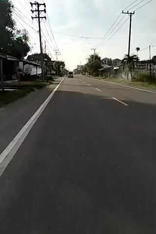 The video shows a static, eye-level view of a road, likely from the side of the road or perhaps a very slow-moving vehicle. The road is paved and features a white line marking the edge of the lane on the left, and a yellow line dividing lanes further ahead. Power lines run overhead, supported by wooden poles on either side of the road. Buildings and trees line the sides of the road. There are a few vehicles visible further down the road. The sky is overcast. The video has no audio.