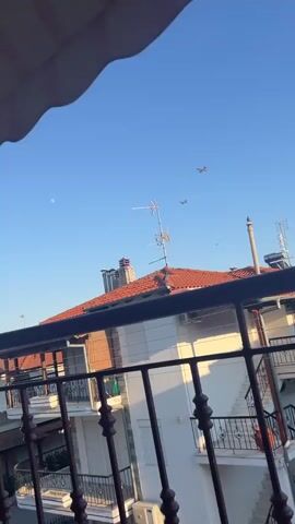 The video shows a clear blue sky above a residential area. The camera is positioned looking upwards from what appears to be a balcony, with a railing in the foreground. On the roof of a building, there is a red tiled roof with chimneys and a television antenna. In the sky, two amphibious aircraft are flying in formation. The aircraft appear to be the same model, with a distinctive high wing and tail configuration. They are seen from a distance against the bright blue sky. The lighting suggests it is daytime, likely late afternoon or early morning given the angle of the light.