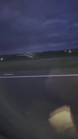 The video, taken from a moving vehicle, shows an airfield at dusk or night. The camera is positioned low, likely inside the vehicle, providing a view of the dark asphalt road ahead, marked with a bright white line. Beyond the road, the airfield stretches out, illuminated by rows of small blue and white lights. In the distance, a line of trees and buildings is faintly visible under a dark, cloudy sky. The vehicle is moving at a moderate speed. Some reflections on the glass of the vehicle obscure the view.