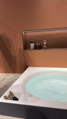 The video opens with a close-up shot of a modern, white jacuzzi bathtub filled with bubbling water and subtle blue light. An integrated cooler on the left side of the tub holds a bottle of champagne and two empty wine glasses. Above the tub, a recessed wooden shelf with lighting displays decorative items, including candles, an hourglass, and small bottles, set against a warm terracotta-colored wall.

The camera then pans slightly to the right, revealing an archway and people moving through a large indoor space. A woman in black trousers and a top walks away from the camera. She is followed by another woman in a dark blue tracksuit with white stripes, carrying a shoulder bag, who walks past, smiling briefly. The view then expands to show a crowd of diverse individuals, both men and women, gathered and walking in a spacious hall. The walls are a consistent terracotta color, and a partial word "smeg" is visible on a distant wall, suggesting a brand presence. The overall scene indicates an exhibition, trade show, or public event taking place indoors, likely in Rho, Italy.
