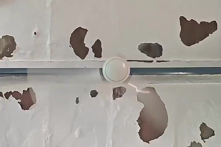 The video shows a white ceiling with peeling paint, revealing brown patches underneath. A horizontal blue pipe runs across the ceiling, and a white circular object, possibly a light fixture or a vent, is mounted centrally on the ceiling, above the pipe. The video is a static shot with no visible people or movement other than the subtle motion blur of the ceiling fan.