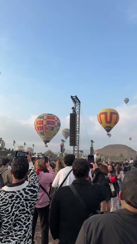 The video captures a scene at what appears to be a festival or outdoor event, with many people looking up at hot air balloons. The balloons are diverse in color and design, some featuring logos like "Sky Balloons," and they float against a clear blue sky with a few wisps of clouds. The crowd is diverse and densely packed, with individuals holding up phones and cameras to capture the scene. A stage-like structure with speakers is visible, hinting at a musical or entertainment element to the event. The ground level is full of people facing toward the balloons.