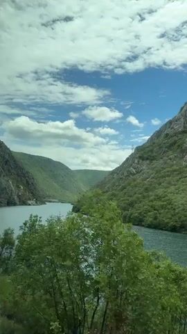 This video captures a serene natural landscape, likely filmed from a moving vehicle given the slight motion blur. The scene is dominated by a wide, tranquil river or lake, its surface a deep, shimmering blue-green. The water is flanked by steep, lush green mountains covered in dense vegetation and interspersed with rugged grey rock formations. The scale of the mountains is imposing, with jagged peaks reaching towards the sky.

The sky above is a vibrant blue, dotted with fluffy white clouds that cast dynamic shadows across the mountainside. The lighting suggests it is daytime, likely late morning or early afternoon, with the sun high enough to illuminate the scene brightly. The overall atmosphere is one of peacefulness and natural beauty.

The camera pans slowly across the vista, offering a panoramic view of the valley and the surrounding geological features. The foreground is partially obscured by the leaves and branches of trees, adding a sense of depth and framing the broader landscape.

While no people or man-made objects are clearly visible, the presence of a road or pathway is implied by the perspective of the filming. The location is identified as Donji Jasenjani, Bosnia and Herzegovina, which places this stunning natural scene in a specific geographical context. The video evokes a feeling of awe and appreciation for the grandeur of the natural world, with no visible text or specific event occurring other than the scenic journey itself.