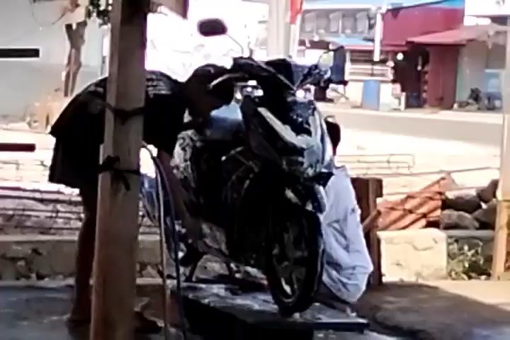 At the 0:00 mark, a dark-colored motorcycle is being washed. Two people are present, one standing and one crouching. The standing person, wearing a black t-shirt and shorts, is on the left side of the motorcycle, facing it and holding onto a wooden post. The crouching person, wearing a white shirt, is on the right side of the motorcycle, facing the rear wheel. The motorcycle is wet and appears to be covered in soap. The environment is outdoors, with a street visible in the background, where another motorcycle is passing by. There are rocks and a building with a red roof in the background. The scene appears to be a motorcycle wash station.