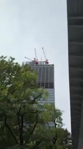 The camera pans across a city scene in Japan. In the background, a tall skyscraper under construction stands out, with two red and white cranes at its peak. A lush green tree partially obscures the view of the building. Moving right, the camera reveals a concrete walkway elevated above street level. People can be seen walking along the street below, which is lined with other buildings and streetlights. The sky is overcast, creating a muted tone for the entire scene. The camera's angle shifts as it passes under the elevated walkway, emphasizing the scale of the construction and urban infrastructure.