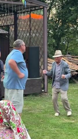 Two men are dancing outdoors in front of a small stage. The man on the left is larger, wearing a blue t-shirt and khaki shorts. He is facing away from the camera. The man on the right is thinner, wearing a grey suit jacket, a grey button-down shirt, khaki pants, and a straw hat. He is facing the camera and appears to be performing a dance routine. A woman in a blue top is visible on the stage behind them, and a woman wearing a brightly colored headscarf is in the foreground, also facing away from the camera. The scene is set in a grassy area with trees and a wooden structure in the background. There are strings of lights and colorful flags hanging from the structure.