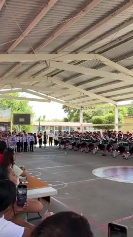 A marching band composed of many young people in uniform stands in formation on a paved, outdoor area under a covered structure. The members of the band are dressed in dark vests with red accents over white shirts, dark pants, and red hats. Most of them are holding drums, and some are holding drumsticks. They appear to be playing. In the background, there are buildings and trees. Some people are visible on the left side of the frame, also in uniform, and some spectators are in the foreground, holding up cell phones to record.